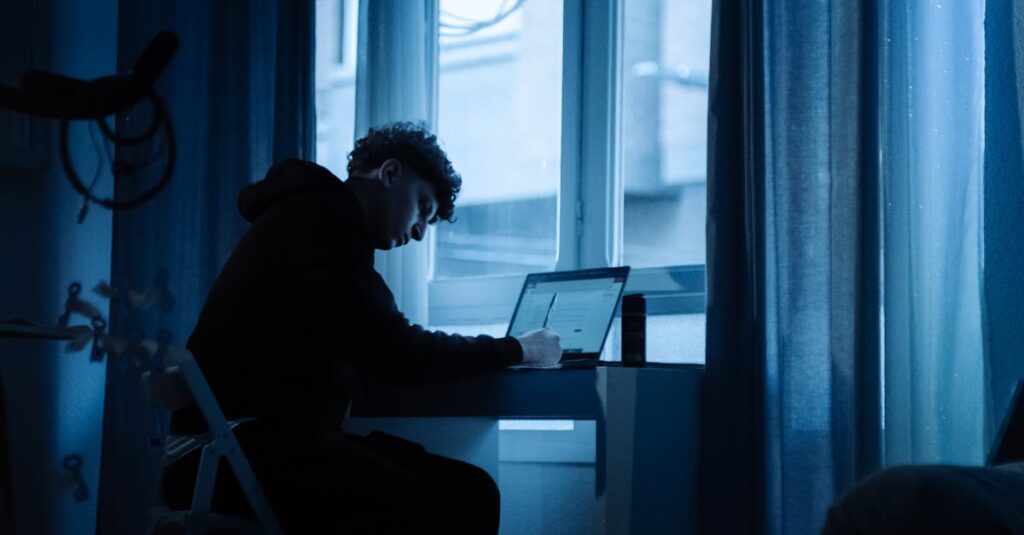 A young man focuses intently on his laptop in a dimly lit room, creating a moody atmosphere.